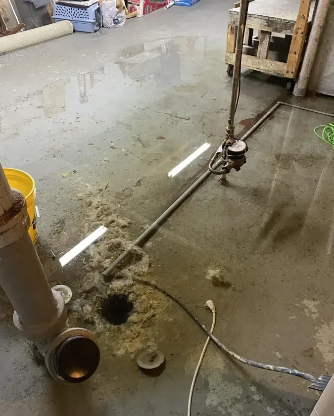 Water and debris surrounding an open drain and exposed plumbing on concrete